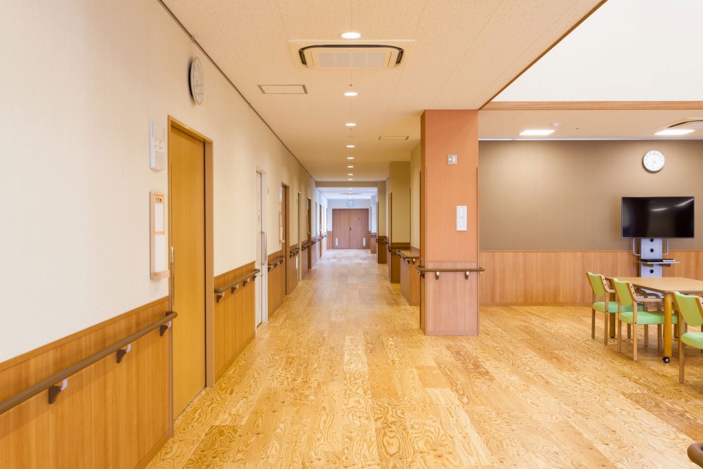 廊下と共用スペースが一体の明るい施設内部。木目調の手摺と床が整備されている。